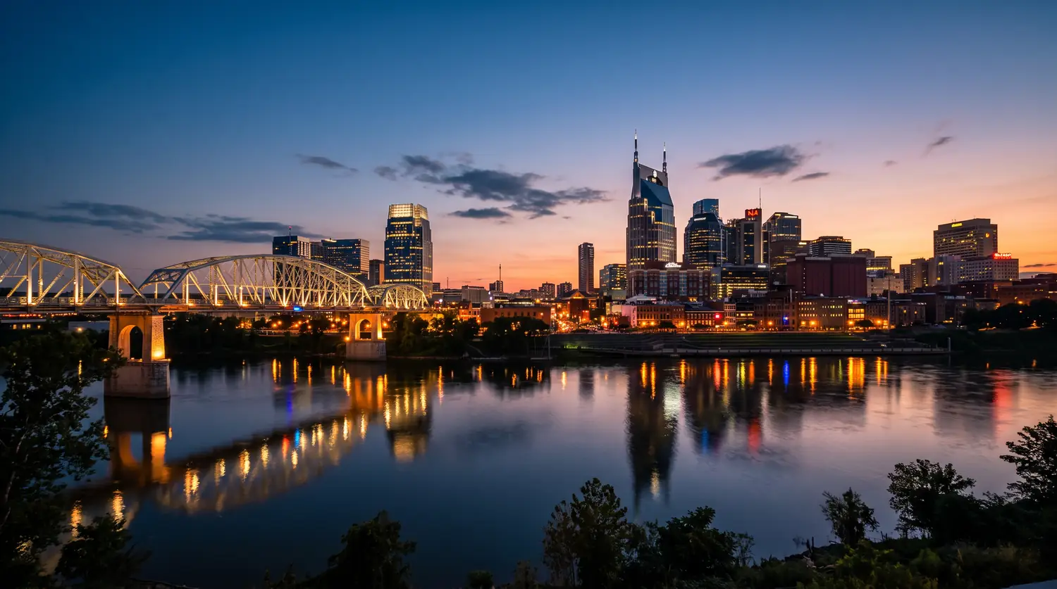 Nashville Skyline at Blue Hour | Music City Wildlife The Nashville skyline at blue-hour dusk beyond the Cumberland River with a calm reflection, a warm sunset gradient in the sky and the lit pedestrian bridge.